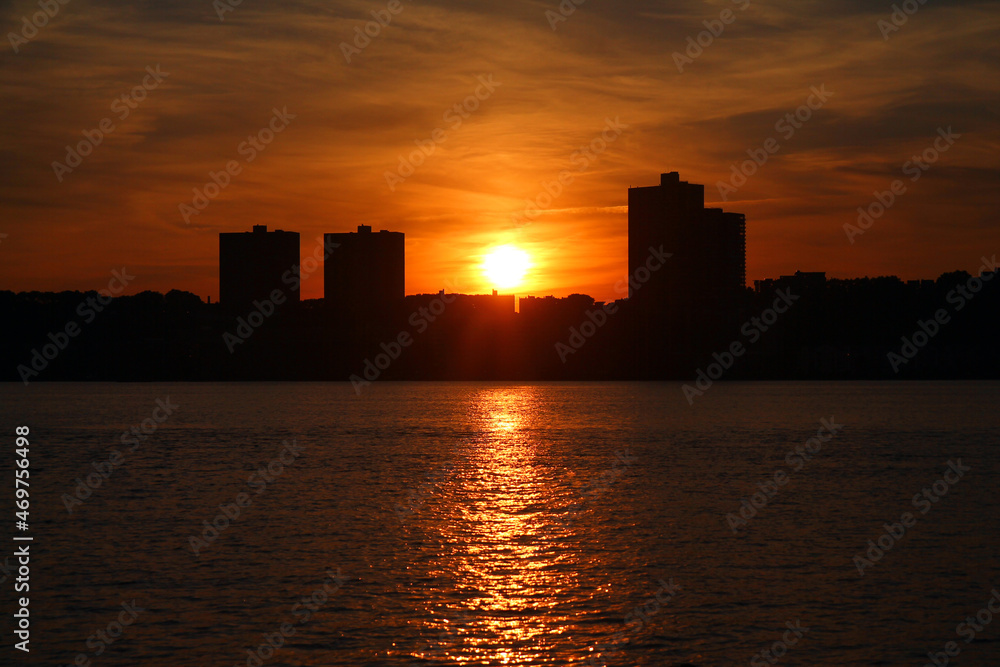 Fototapeta premium The orange and round sun going behind the skyscrapers and illuminating the river