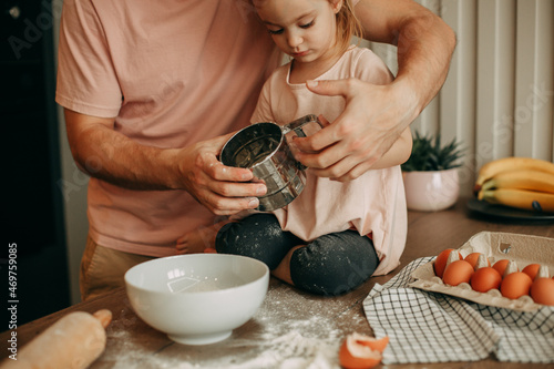 Dad prepares dough with his daughter at home in the kitchen. A young man in a pink T-shirt helps his daughter with light brown hair and a pink dress and gray pants to hold a sieve with flour