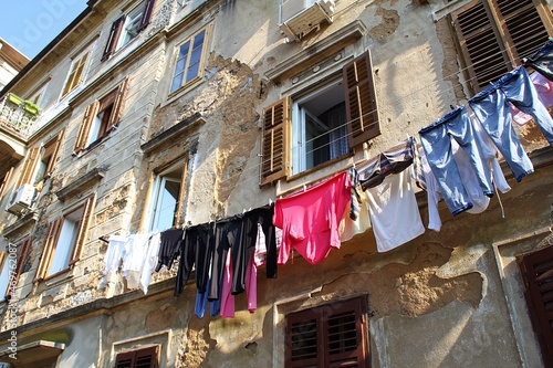 old, crumbling wall, open window in an old building, laundry hanging on a string, falling plaster, old building, poor district, dilapidated walls, colorful laundry, old shutters © AMTM