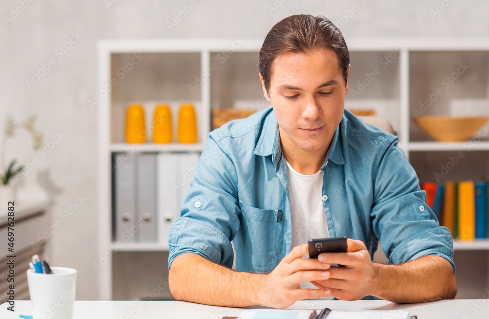 Young man with a smartphone sits at the table at home. Freelancer, remote worker or student workplace. Distant job concept. Young man with a smartphone sits at the table at home. Freelancer, remote worker or student workplace. Distant job concept.