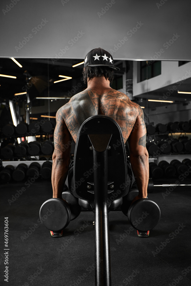 Fototapeta Strong and muscular dark skin man trains on modern equipment in gym. Portrait of muscular pumped up fitness trainer