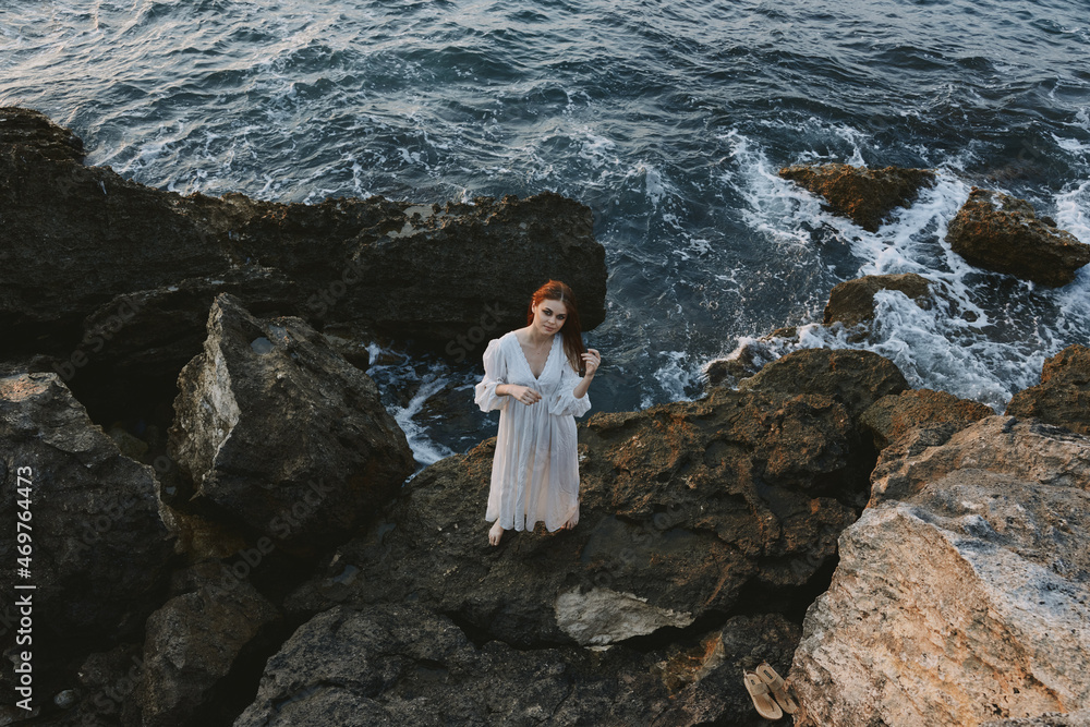 A woman in a white dress stands barefoot on the stones on the ocean shore top view