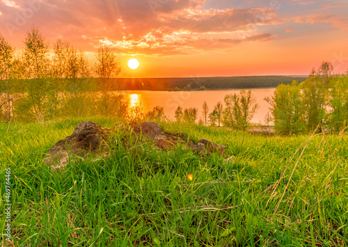 Scenic view at beautiful sunset on a shiny lake with old rough stone on the foreground, green grass, birch trees, golden sun rays, calm water ,nice cloudy sky on a background, spring landscape