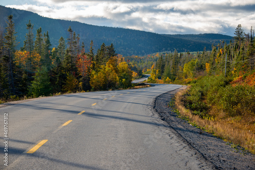 A single broken yellow line divides a two lane paved highway. The road is dry pavement and on both sides are trees, shrubs, and pants with autumn colors. The road is curved with blue sky and clouds.