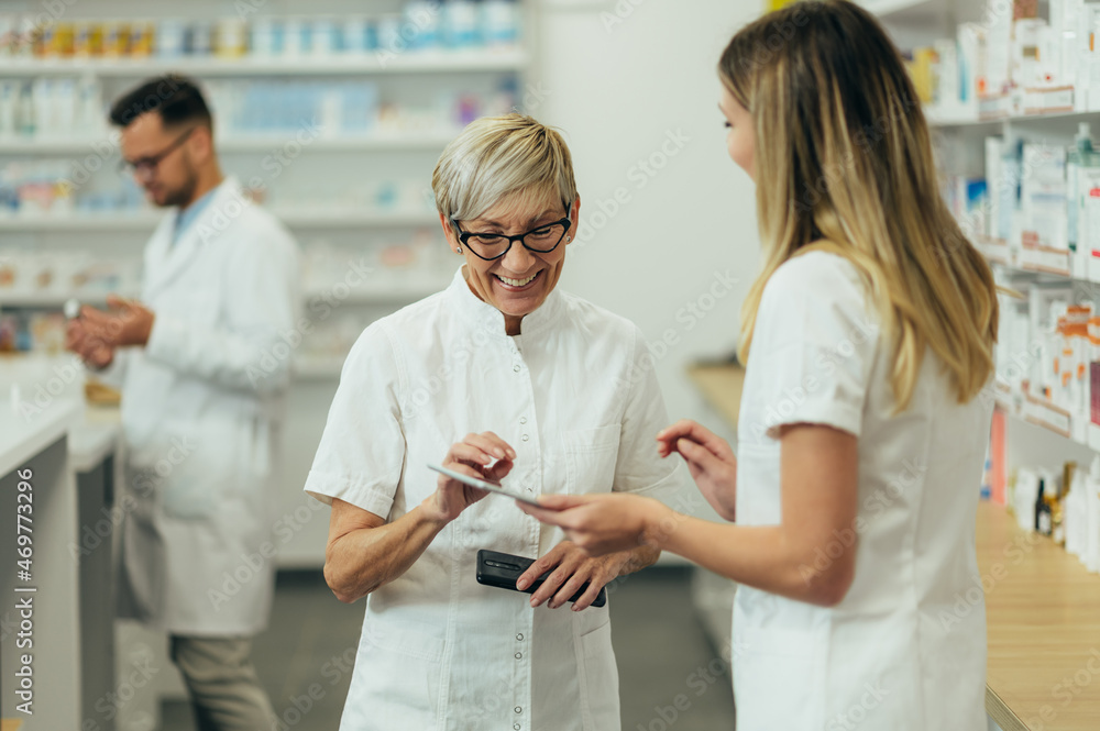 Senior female pharmacist using a tablet while working in a pharmacy with her young colleague Senior female pharmacist using a tablet while working in a pharmacy with her young colleague