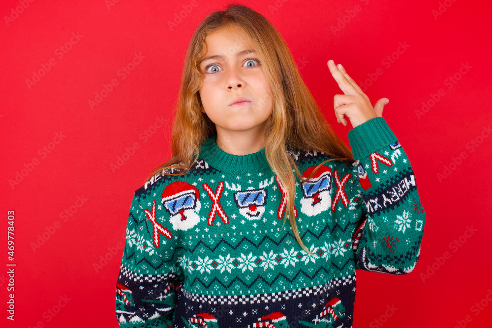 Unhappy little kid girl wearing knitted sweater christmas over red background imitates gun shoot makes suicide gesture keeps two fingers on temples. Unhappy little kid girl wearing knitted sweater christmas over red background imitates gun shoot makes suicide gesture keeps two fingers on temples.