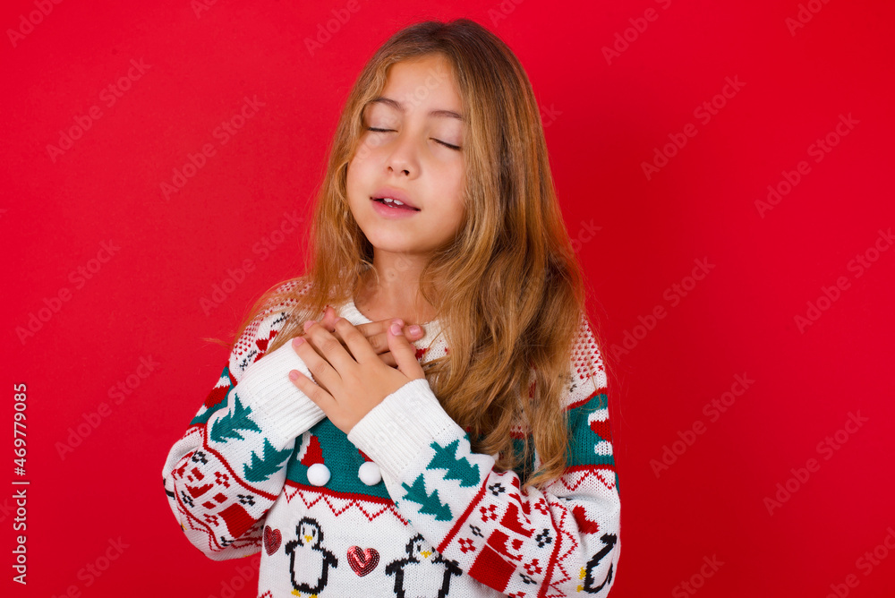 little kid girl wearing knitted sweater christmas over red background expresses happines, laughs pleasantly, keeps hands on heart little kid girl wearing knitted sweater christmas over red background expresses happines, laughs pleasantly, keeps hands on heart