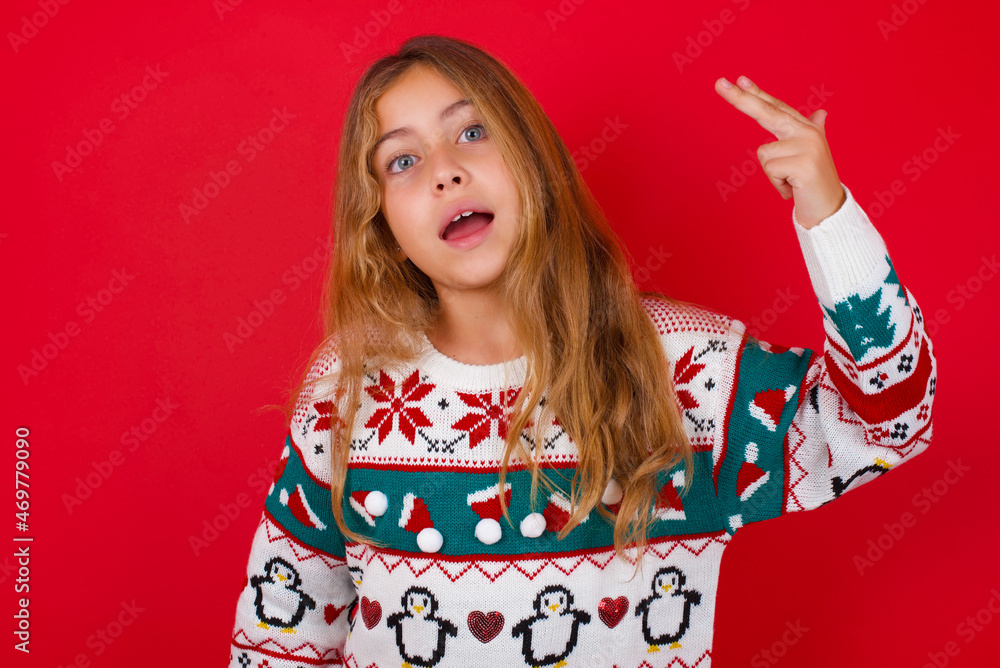 little kid girl wearing knitted sweater christmas over red background foolishes around shoots in temple with fingers makes suicide gesture. Funny model makes finger gun pistol little kid girl wearing knitted sweater christmas over red background foolishes around shoots in temple with fingers makes suicide gesture. Funny model makes finger gun pistol
