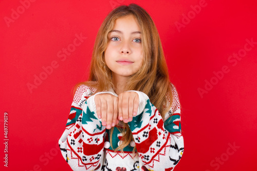 little kid girl wearing knitted sweater christmas over red background makes bunny paws and looks with innocent expression plays with her little kid