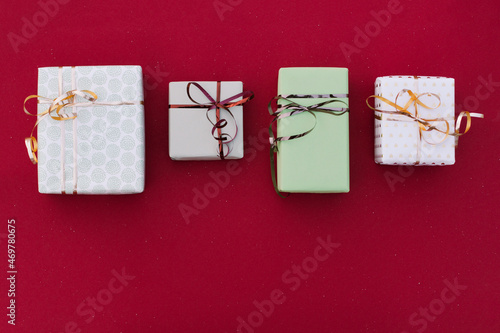 four boxes of gifts in gray, green and white wrapping paper with golden hearts tied with shiny ribbons with bows lie on a red background, visible on a red background strewn with snowflakes