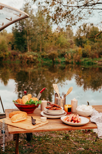 table set near trailer of mobile home or recreational vehicle stands on shore of pond in camping in autumn, concept of family local travel in native country on caravan or camper van and camping life