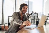 Young man host in headphones and glasses enjoying podcasting in studio, speaking into a microphone, holding a pen, using laptop. Handsome podcaster laughing while streaming live audio podcast