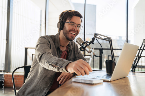 Young man host in headphones and glasses enjoying podcasting in studio, speaking into a microphone, holding a pen, using laptop. Handsome podcaster laughing while streaming live audio podcast photo