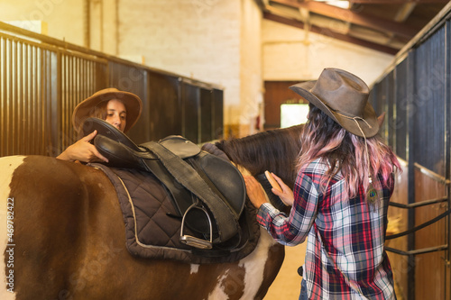 Two cowgirl women preparing to ride a horse in a stable, southern usa hats and jeans, vertical photo
