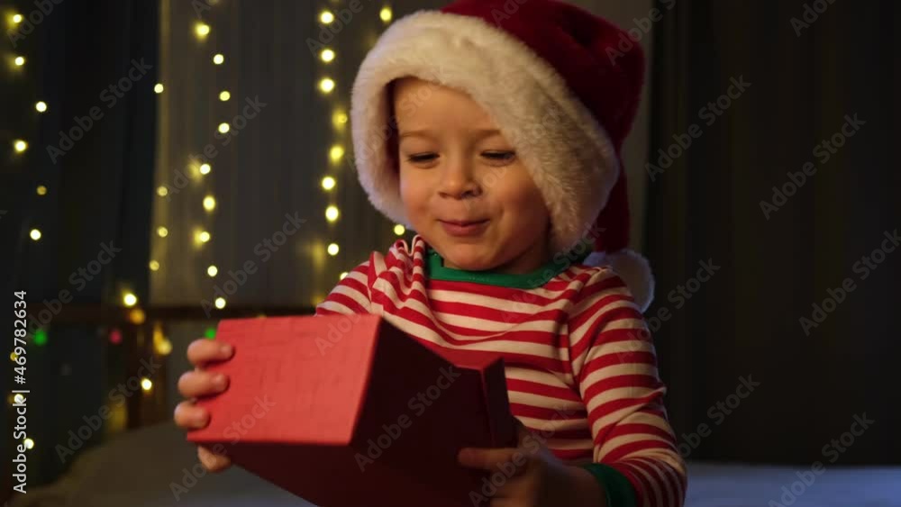Christmas boy jumping in Santa hat at home evening. New Year and Holidays. Christmas happiness. The boy in the Santa Claus hat having fun, enjoying Christmas. Festive atmosphere around the child. Christmas boy jumping in Santa hat at home evening. New Year and Holidays. Christmas happiness. The boy in the Santa Claus hat having fun, enjoying Christmas. Festive atmosphere around the child.
