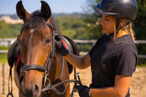 Caucasian blonde girl on a horse stroking and pampering a brown horse, dressed in black rider with safety hat