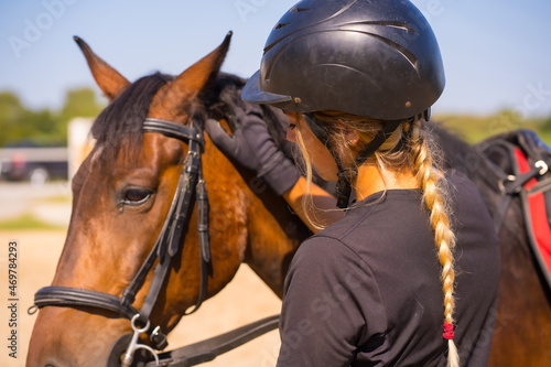 Caucasian blonde girl on a horse stroking and pampering a brown horse, dressed in black rider with safety hat © unai