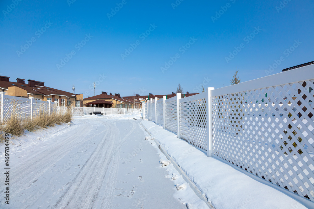 Snowy road in a cottage village. White plastic fence in a modern cottage village on a clear winter day. Snow drifts in front of a vinyl fence against a blue sky