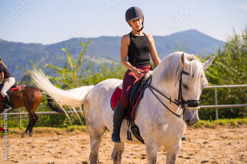 Caucasian blonde girl riding a white horse, dressed in black rider with safety hat © unai
