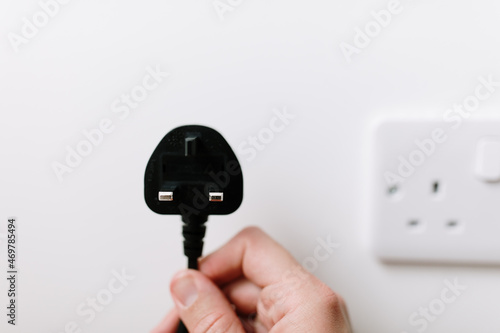 A hand is holding a black plug from the United Kingdom. Unplugged concept due to high energy prices. A tip for saving energy. A blurry socket in the background. White wall, natural light. Copy space.