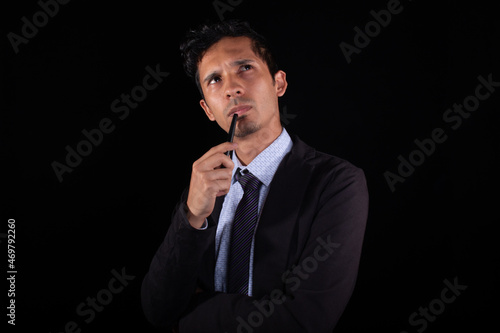 Thoughtful businessman looking up and placing a pen in his mouth, isolated on black background. Young adult latin man thinking about solution.