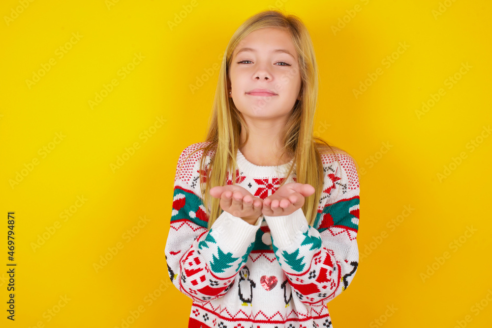 caucasian little kid girl wearing knitted sweater christmas over yellow background holding something with open palms, offering to the camera. caucasian little kid girl wearing knitted sweater christmas over yellow background holding something with open palms, offering to the camera.