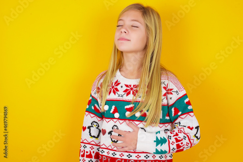 caucasian little kid girl wearing knitted sweater christmas over yellow background touches tummy, smiles gently, eating and satisfaction concept.