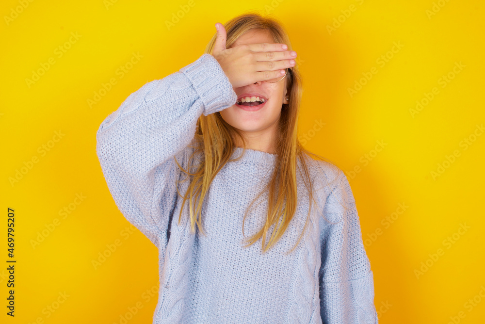caucasian kid girl wearing blue knitted sweater over yellow background smiling and laughing with hand on face covering eyes for surprise. Blind concept. caucasian kid girl wearing blue knitted sweater over yellow background smiling and laughing with hand on face covering eyes for surprise. Blind concept.