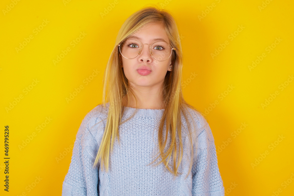 Shot of pleasant looking caucasian kid girl wearing blue knitted sweater over yellow background , pouts lips, looks at camera, Human facial expressions Shot of pleasant looking caucasian kid girl wearing blue knitted sweater over yellow background , pouts lips, looks at camera, Human facial expressions