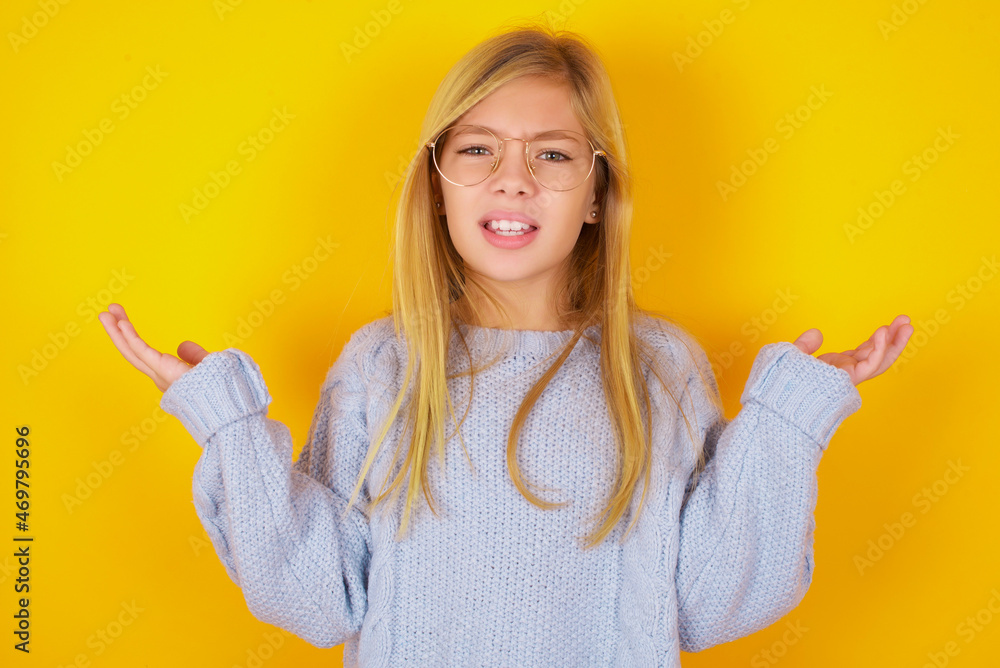 Indignant caucasian kid girl wearing blue knitted sweater over yellow background gestures in bewilderment, frowns face with dissatisfaction. Indignant caucasian kid girl wearing blue knitted sweater over yellow background gestures in bewilderment, frowns face with dissatisfaction.