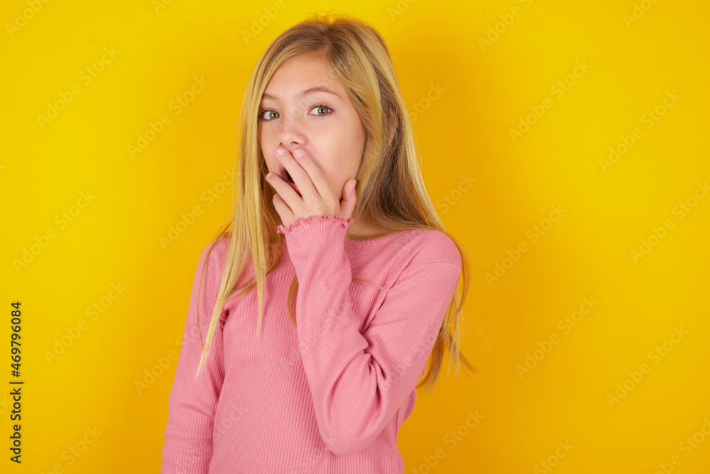 caucasian little kid girl wearing long sleeve shirt over yellow background covers mouth and looks with wonder at camera, cannot believe unexpected rumors. caucasian little kid girl wearing long sleeve shirt over yellow background covers mouth and looks with wonder at camera, cannot believe unexpected rumors.