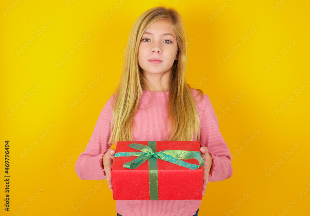caucasian little kid girl wearing long sleeve shirt over yellow background holding a present. caucasian little kid girl wearing long sleeve shirt over yellow background holding a present.