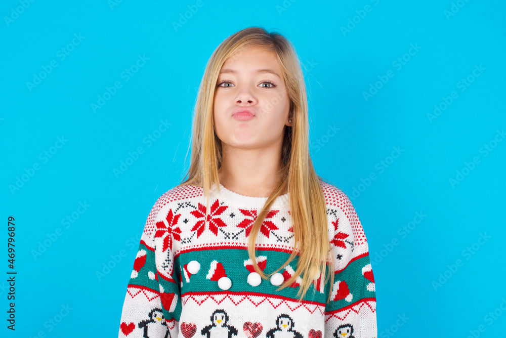 caucasian little kid girl wearing knitted sweater christmas over blue background crosses eyes, puts lips, makes grimace with awkward expression has fun alone, plays fool. caucasian little kid girl wearing knitted sweater christmas over blue background crosses eyes, puts lips, makes grimace with awkward expression has fun alone, plays fool.