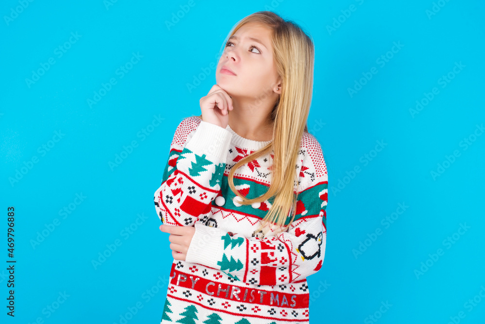 Portrait of thoughtful caucasian little kid girl wearing knitted sweater christmas over blue background keeps hand under chin, looks away trying to remember something or listens something Portrait of thoughtful caucasian little kid girl wearing knitted sweater christmas over blue background keeps hand under chin, looks away trying to remember something or listens something