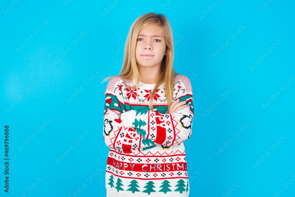 caucasian little kid girl wearing knitted sweater christmas over blue background bitting his mouth and looking worried and scared crossing arms, worry and doubt. caucasian little kid girl wearing knitted sweater christmas over blue background bitting his mouth and looking worried and scared crossing arms, worry and doubt.