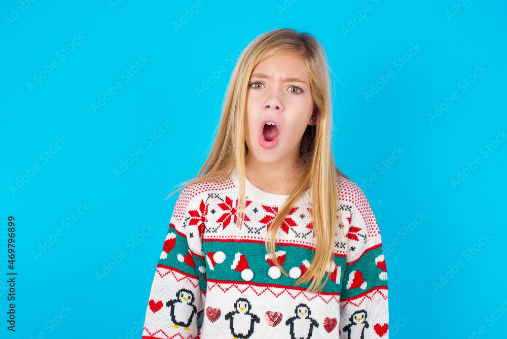 Expressive facial expressions. Shocked stupefied caucasian little kid girl wearing knitted sweater christmas over blue background , keeps jaw dropped feels stunned from what he sees aside. Expressive facial expressions. Shocked stupefied caucasian little kid girl wearing knitted sweater christmas over blue background , keeps jaw dropped feels stunned from what he sees aside.
