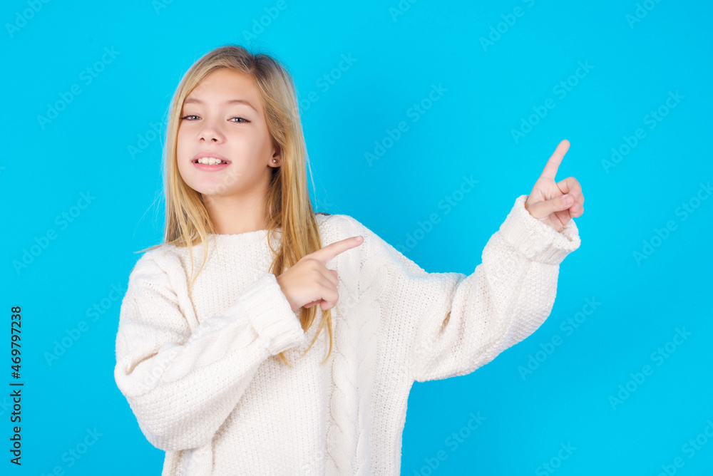 Positive caucasian little kid girl wearing wool jersey over blue background with beaming smile pointing with two fingers and looking on empty copy space. Advertisement concept. Positive caucasian little kid girl wearing wool jersey over blue background with beaming smile pointing with two fingers and looking on empty copy space. Advertisement concept.