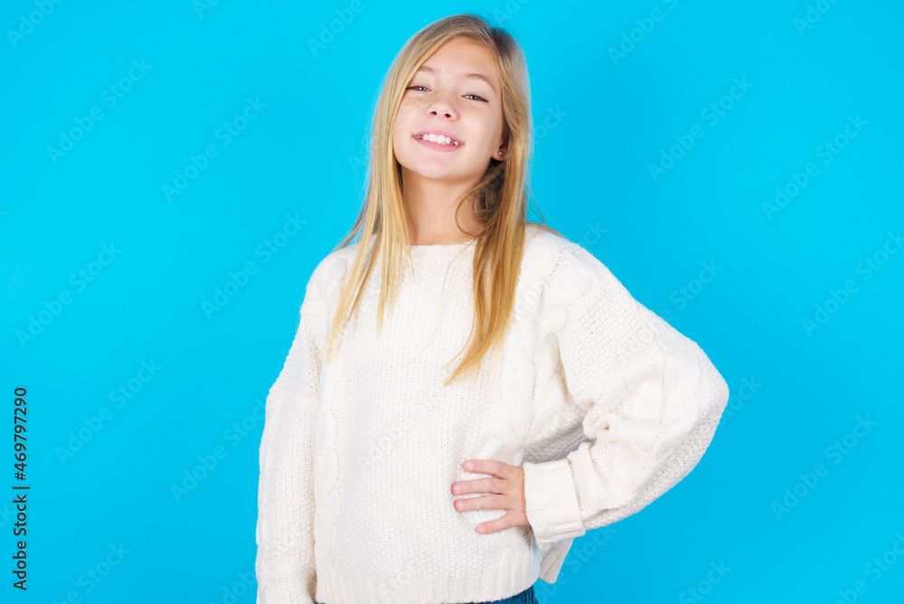 Studio shot of cheerful caucasian little kid girl wearing wool jersey over blue background keeps hand on hip, smiles broadly. Studio shot of cheerful caucasian little kid girl wearing wool jersey over blue background keeps hand on hip, smiles broadly.