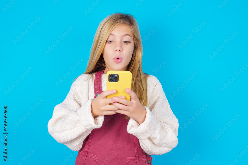 caucasian little kid girl wearing jumpsuit over blue background looks with bugged eyes, holds modern smart phone, receives unexpected message from friend, reads reminder. caucasian little kid girl wearing jumpsuit over blue background looks with bugged eyes, holds modern smart phone, receives unexpected message from friend, reads reminder.