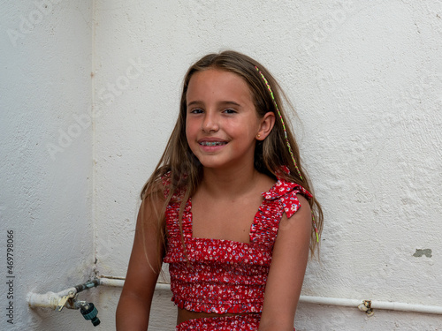 A photo of a young caucasian girl in red dress sitting and smiling next to an old pipe on a white wall background
