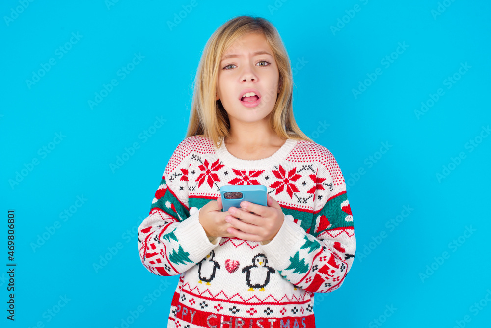 Photo of astonished crazy caucasian little kid girl wearing knitted sweater christmas over blue background hold smartphone dislike feedback concept Photo of astonished crazy caucasian little kid girl wearing knitted sweater christmas over blue background hold smartphone dislike feedback concept