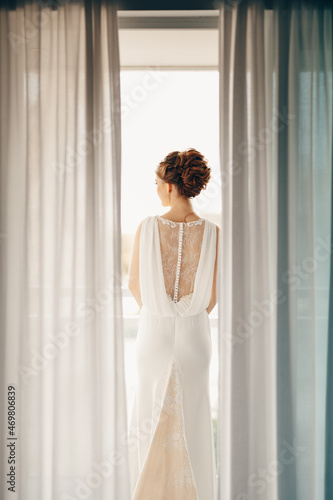 Bride in a white lace dress with a beautiful high hairdo stands on the balcony. View from the room