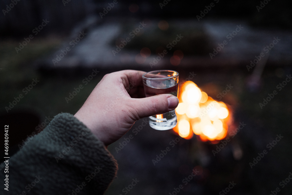 Draußen am Lagerfeuer Schnaps trinken. Mann hält Schnapsglas vor einem Feuer Draußen am Lagerfeuer Schnaps trinken. Mann hält Schnapsglas vor einem Feuer