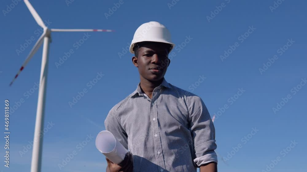 African American electrical engineer in a helmet and with a drawing in his hand stands on the background of a windmill at an air power plant African American electrical engineer in a helmet and with a drawing in his hand stands on the background of a windmill at an air power plant