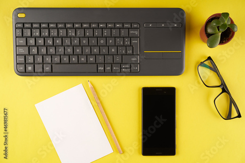 top view yellow desk with keyboard, glasses, notepad, pencil, plant and cell phone