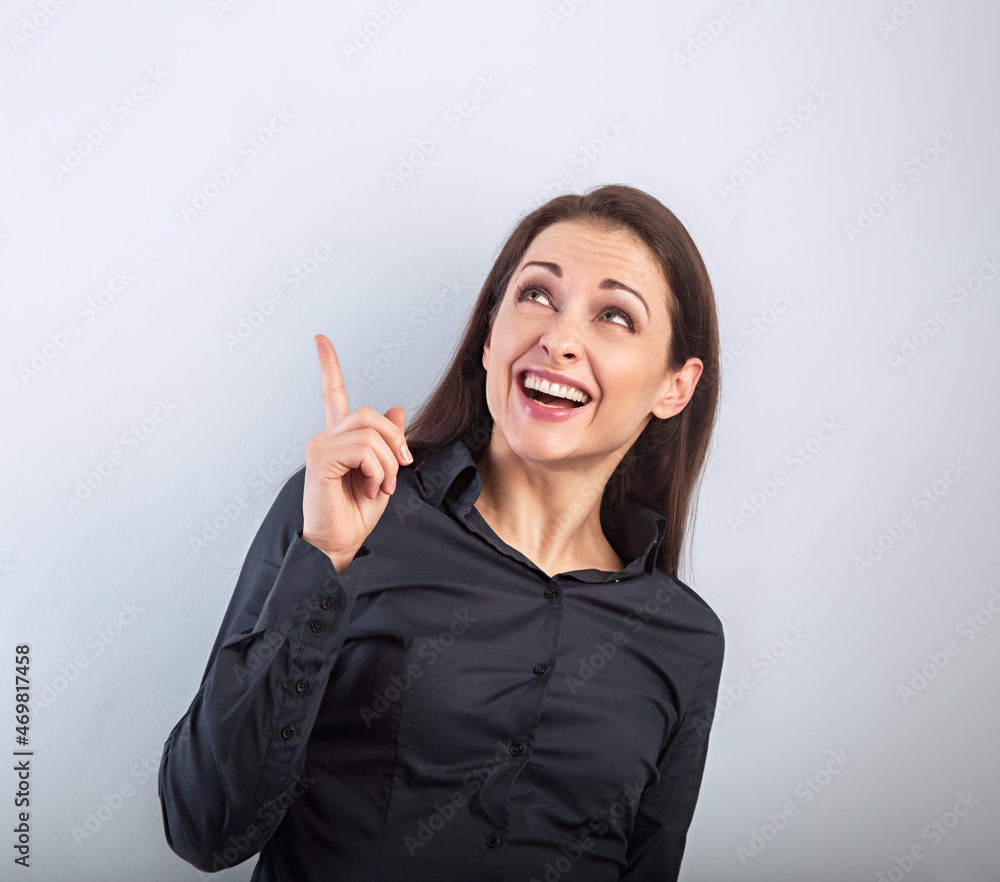 Beautiful excited business woman in black t-shirt pointing the finger up with toothy smiling on empty copy space blue studio background. Closeup Beautiful excited business woman in black t-shirt pointing the finger up with toothy smiling on empty copy space blue studio background. Closeup