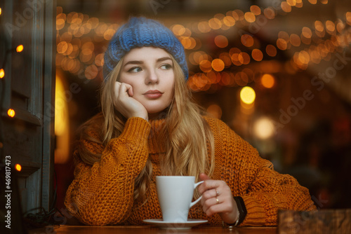 Cute thoughtful woman holding cup of coffee or tea, sitting, posing at cafe. Winter vacation, Christmas holidays conception. Model wearing knitted beanie hat, sweater. Copy, empty space for text photo