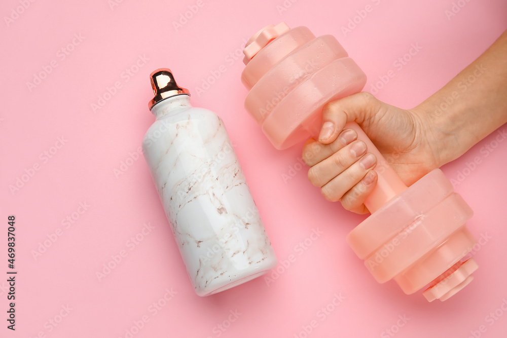 Fototapeta premium Female hand with dumbbell and bottle of water on pink background