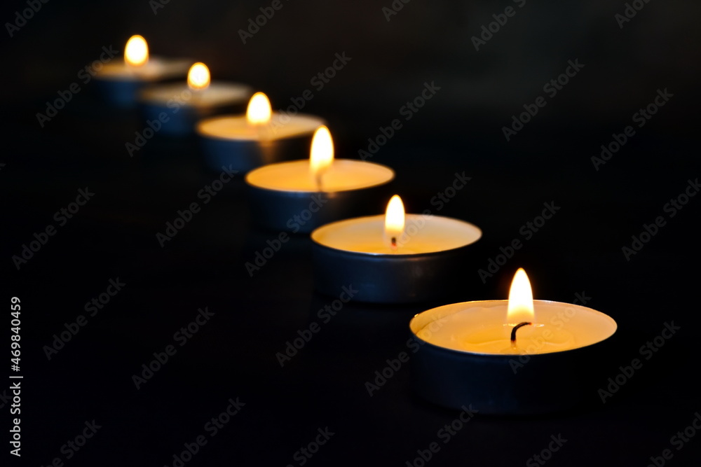 Lit decorative tea candles close up placed in a row with a blurred background in the dark. Selective focus on first candle Lit decorative tea candles close up placed in a row with a blurred background in the dark. Selective focus on first candle