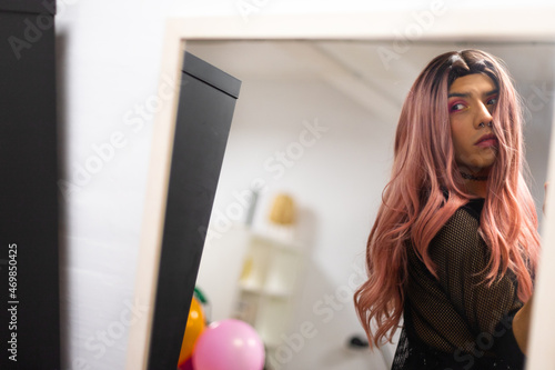 Young caucasian man wearing long pink wig and makeup looking at himself in the mirror. Copy space.